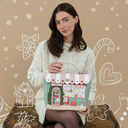 A woman wearing a cream knit sweater holds a decorative handbag styled like a candy store. The bag has a pink and white striped awning, a mint green body, and various candy-themed illustrations, set against a beige background with white holiday doodles.