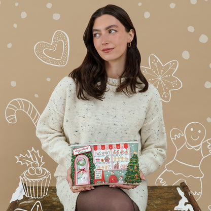 A woman wearing a cream knit sweater holds a Christmas-themed advent calendar. The calendar features festive illustrations including a Christmas tree, candy canes, a gingerbread man, and a snowman door, with the text 'Merry Christmas' at the top.