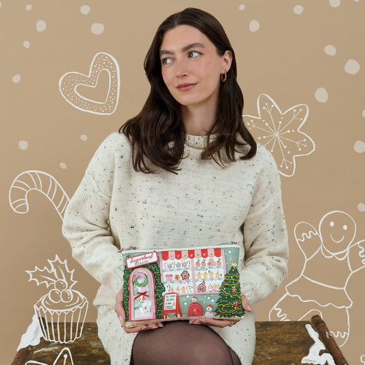 A woman wearing a cream knit sweater holds a Christmas-themed advent calendar. The calendar features festive illustrations including a Christmas tree, candy canes, a gingerbread man, and a snowman door, with the text 'Merry Christmas' at the top.