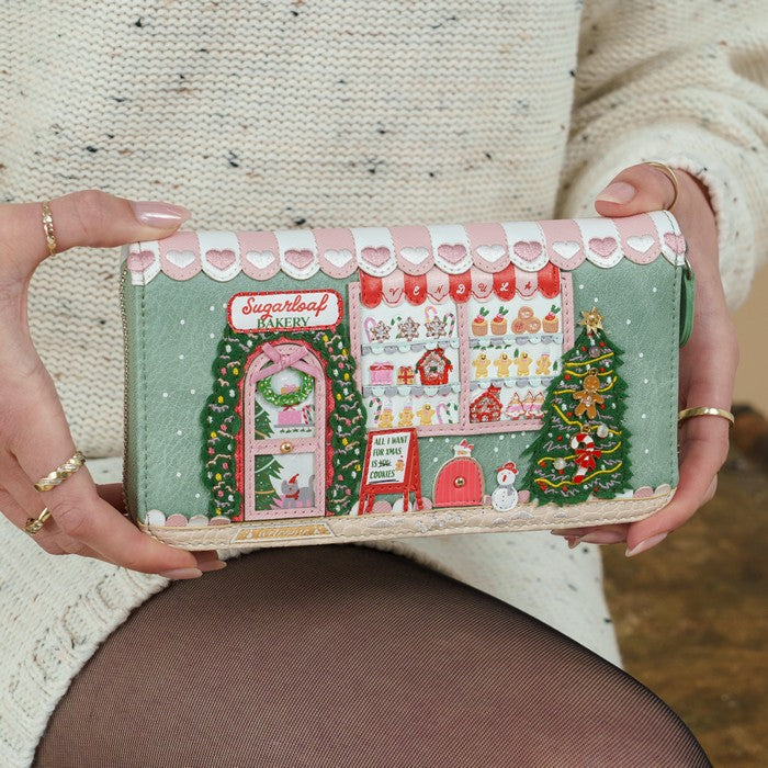 A person holds a long wallet or clutch featuring a detailed Christmas bakery storefront illustration with pink hearts, green trees, and holiday decorations. The person is wearing a cream knit sweater and dark tights.