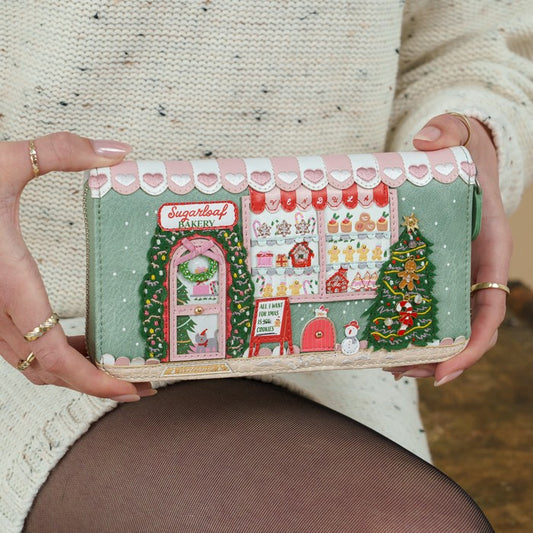 A person holds a long wallet or clutch featuring a detailed Christmas bakery storefront illustration with pink hearts, green trees, and holiday decorations. The person is wearing a cream knit sweater and dark tights.