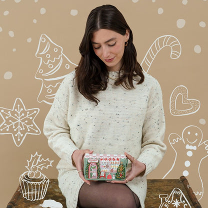 A woman in a cream knit sweater holds a small gingerbread-themed advent calendar against a warm beige background decorated with white holiday illustrations.