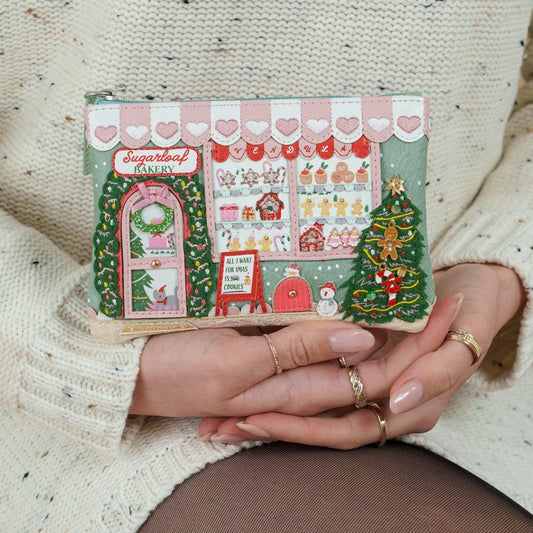 A person in a cream knit sweater holds a small rectangular fabric pouch or wallet featuring a whimsical bakery storefront design with pink hearts, a decorated Christmas tree, and holiday illustrations.