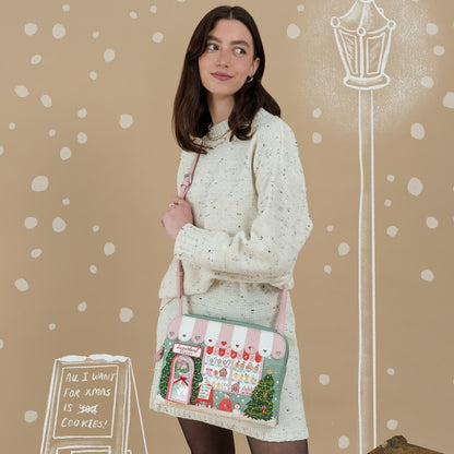 A woman wearing a cream knit sweater and dress stands against a beige holiday-themed backdrop, holding a small shoulder bag decorated with a Christmas market scene, pink striped awning, and a Christmas tree.