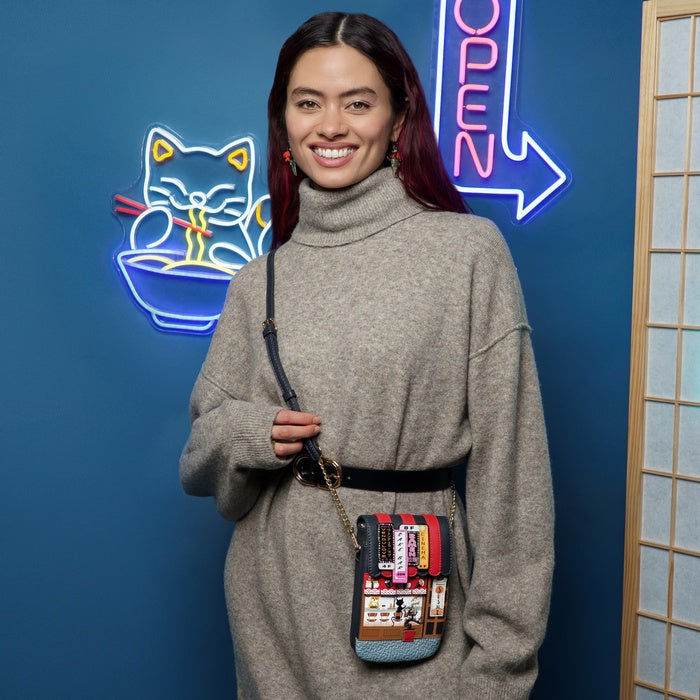 Woman holding a colorful crossbody bag in front of a blue wall with neon signs.