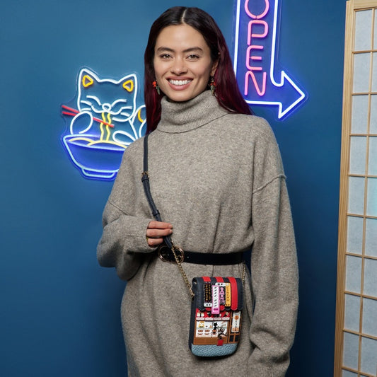 Woman holding a colorful crossbody bag in front of a blue wall with neon signs.