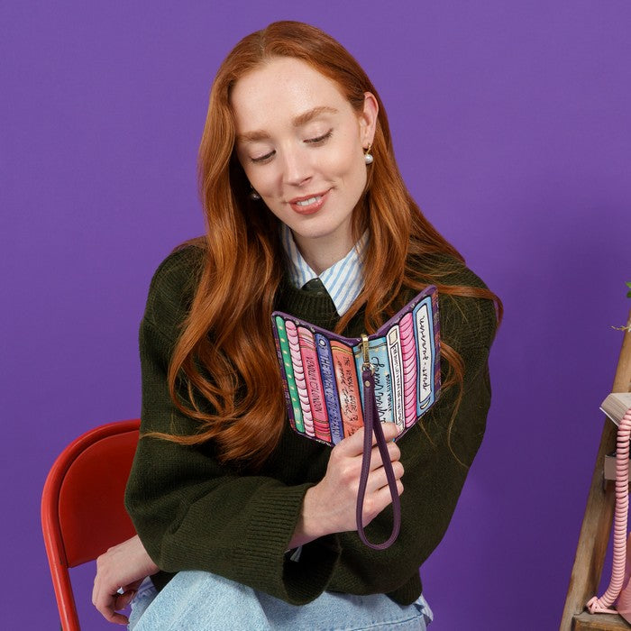 A woman with long red hair sits against a purple background holding an open zip-around card holder wallet decorated with colorful book spines. She is wearing a dark green knit sweater, a striped collared shirt, and light blue jeans.
