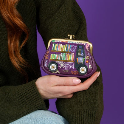 A person in a dark green knit sweater holds a small purple kiss-lock purse shaped like a bookshelf bus with colorful book spines and illustrated details, against a solid purple background.