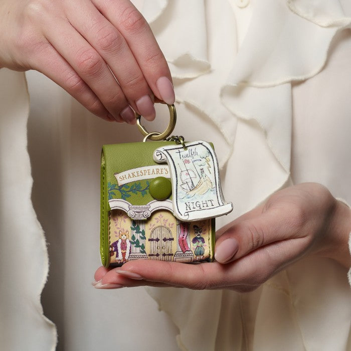 Hand holding a decorative keychain with a miniature book design against a neutral background