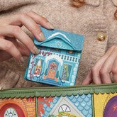A woman with curly red hair sits on a green velvet sofa, smiling as she opens a colorful zippered clutch bag. The clutch features a vibrant, illustrated storefront design with the text 'Piazza'.