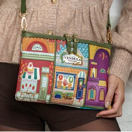 A person wearing a beige knit sweater holds a small crossbody bag with a colorful illustrated storefront pattern featuring purple, red, green, and blue buildings.