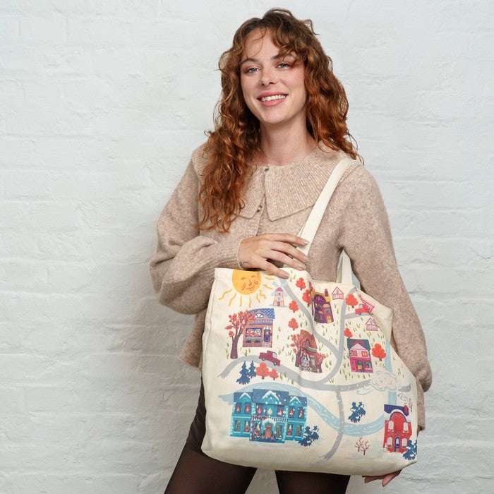 A smiling woman with curly red hair holds a large cream canvas tote bag featuring a colorful winter village illustration with houses, trees, and a sun, posed against a white brick wall.