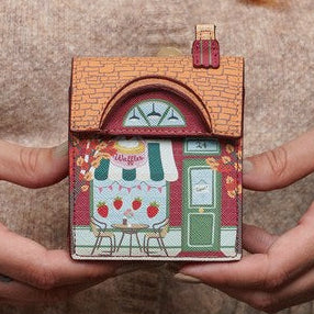 A person in a beige knit sweater holds a small decorative house-shaped item between both hands. The item has a brown roof, a white body, red accents, and a green door.