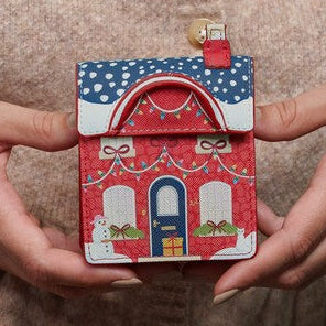 A person in a beige knit sweater holds a small decorative house-shaped coin purse or card holder with a red roof, blue polka-dot top, and white trim. The scene includes hands, part of the torso, knitting needles, and yarn in a cozy indoor setting.