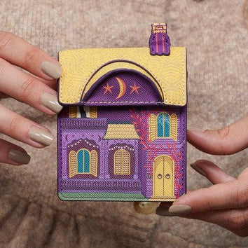 A person in a beige knit sweater holds a small house-shaped accessory with a yellow roof and purple body, featuring embroidered details like a crescent moon and star.