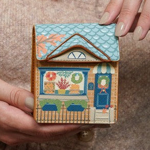 A person in a beige knit sweater holds a small handcrafted wooden house ornament with a colorful painted window and blue roof, while another hand adjusts it. The background is softly blurred, showing a teal garment.