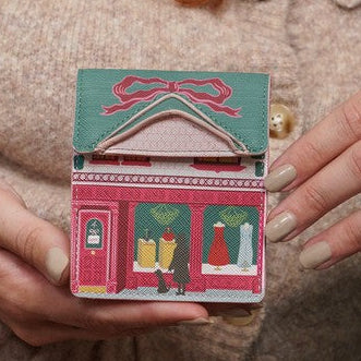A close-up of a person holding a small, colorful, house-shaped card or ornament with a pink storefront design, wearing a beige knit cardigan.