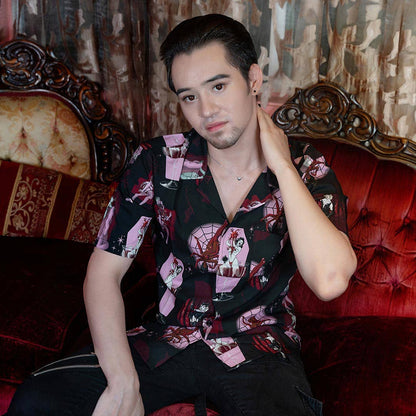 A man wearing a black short-sleeve button-up shirt with a pink, red, and white graphic print, sitting on a red velvet tufted sofa in an ornate room.