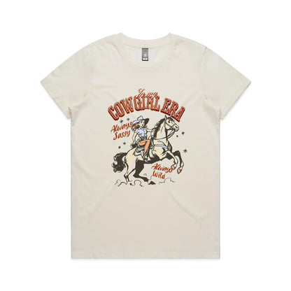 A white graphic t-shirt featuring a retro western illustration of a cowgirl riding a horse. The text reads 'In my COWGIRL ERA' in arched red letters, 'Always Sassy' in red script, and 'Always Wild' in red script, with the cowgirl wearing a cowboy hat and boots while riding a rearing horse.
