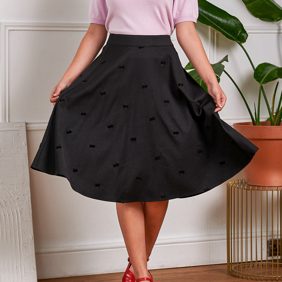 Person wearing a black skirt with heart patterns in a room with a plant and decorative items.