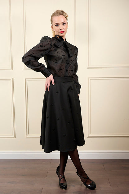 Woman wearing a black outfit with a high collar and a long skirt against a beige paneled wall.