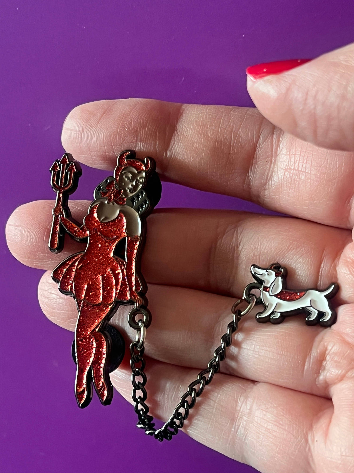 A hand holding a black metal chain accessory with two charms: a red glitter pin featuring a woman in a dress and a small white dog charm with a red heart accent, set against a solid purple background.