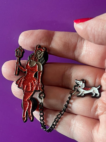 A hand holding a black metal chain accessory with two charms: a red glitter pin featuring a woman in a dress and a small white dog charm with a red heart accent, set against a solid purple background.