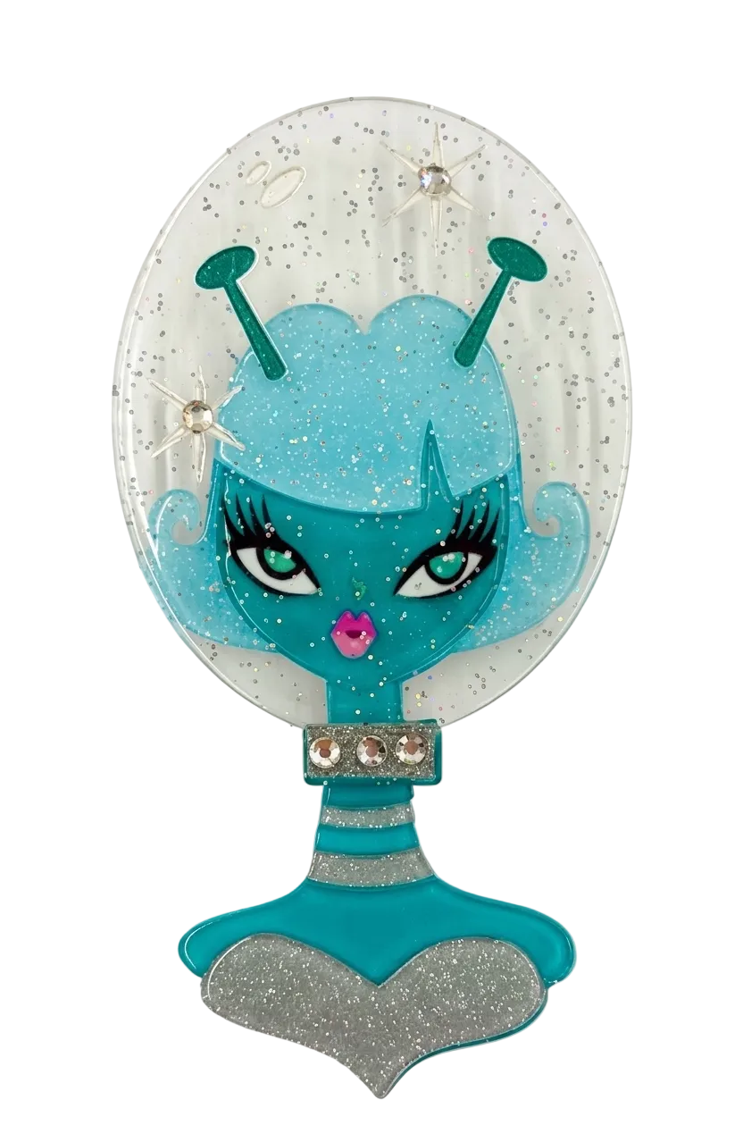 A decorative oval mirror with a glittery white reflective surface featuring a cartoon alien face in teal and light blue, with rhinestone accents and a silver heart-shaped base.