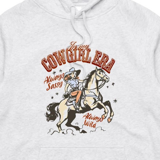 A close-up of a light heather gray hoodie featuring a vintage-style graphic of a cowboy riding a horse, with the text 'In my COWGIRL ERA Always Sassy Always Wild'.