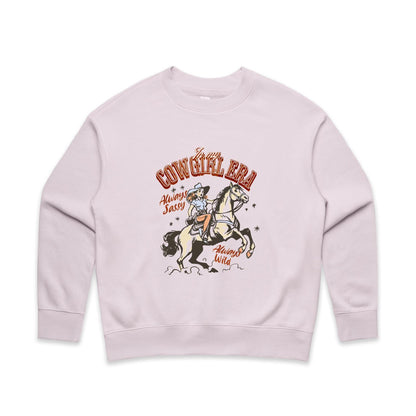 A close-up of a cream-colored t-shirt featuring a vintage-style western graphic. The graphic shows a cowboy riding a rearing horse, with retro text reading 'In my COWGIRL ERA', 'Always Sassy', and 'Always Wild' in reddish-brown and black.