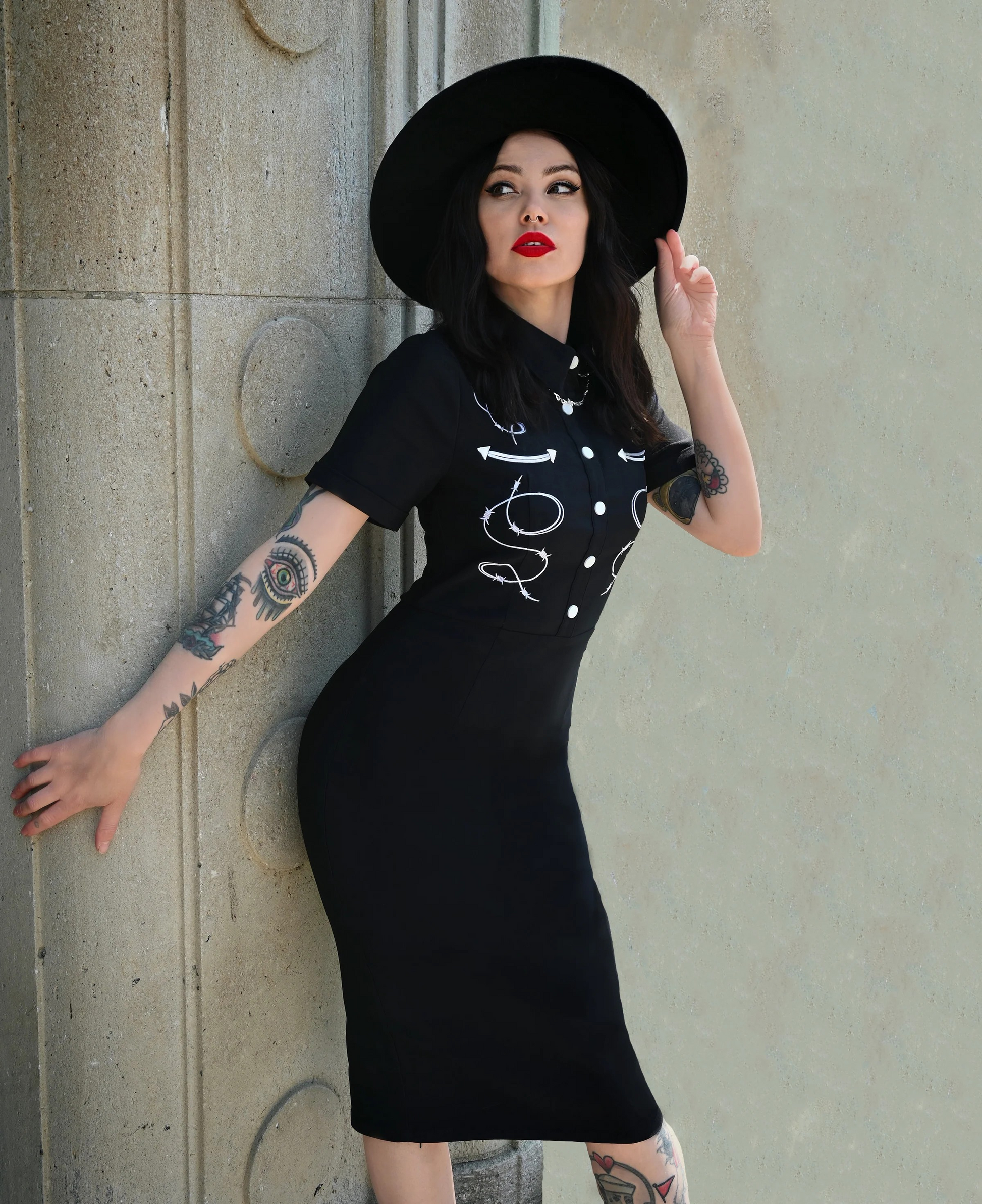 Woman wearing a black dress with white designs and a black hat against a concrete wall.
