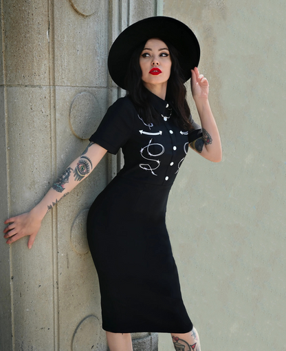 Woman wearing a black dress with white designs and a black hat against a concrete wall.