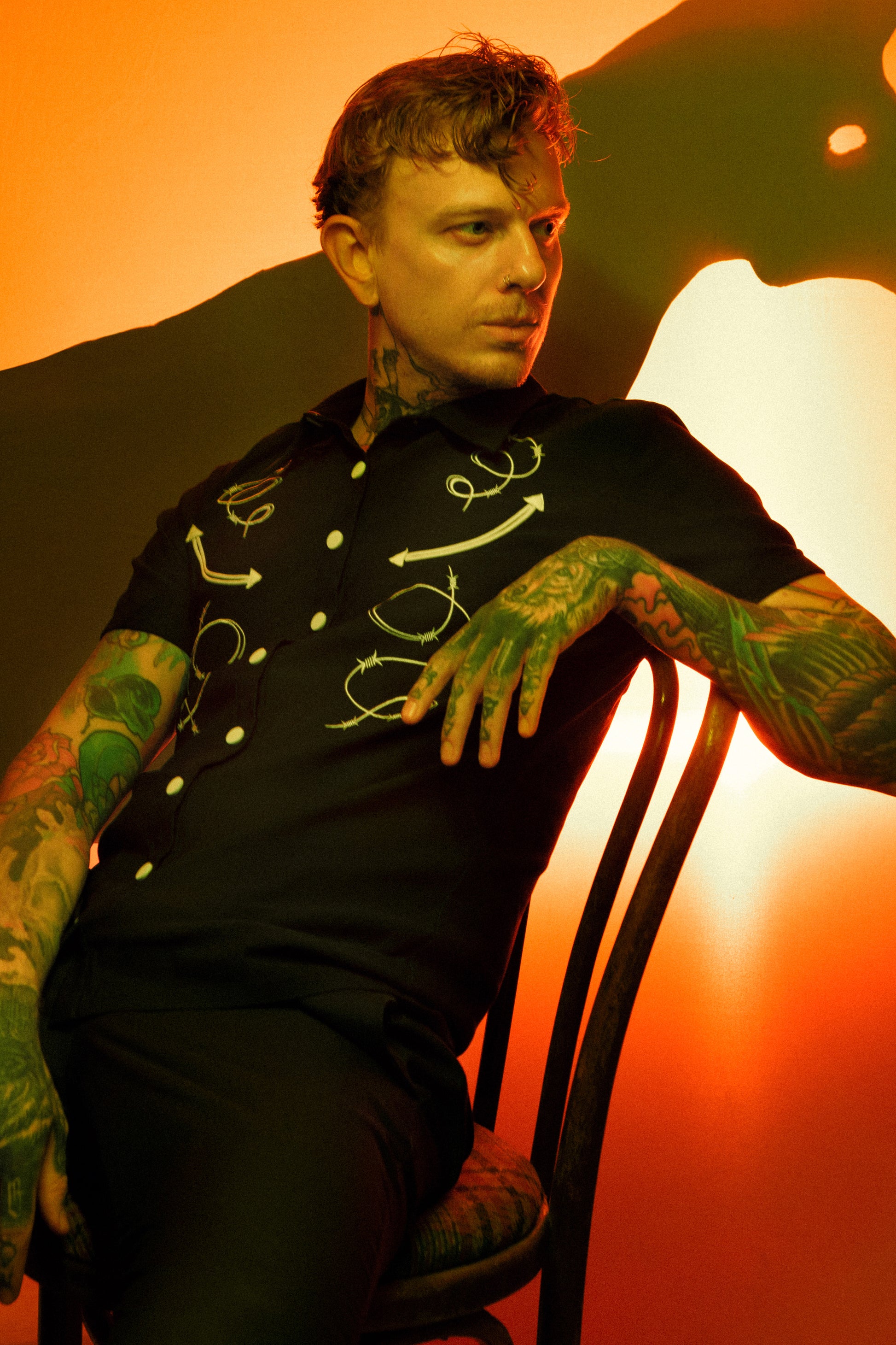 Man with tattoos sitting on a chair against an orange background