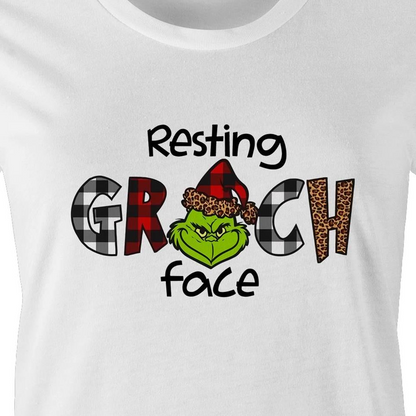 White crewneck t-shirt featuring a graphic print of the Grinch wearing a red and black plaid Santa hat, with the text 'Resting GRCH face' in black.