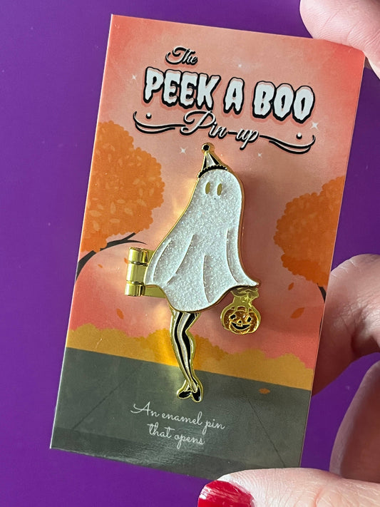 A hand holds a Halloween-themed enamel pin on its backing card. The pin is a ghost character with a glittery white body, gold trim, and a small gold jack-o'-lantern bag.
