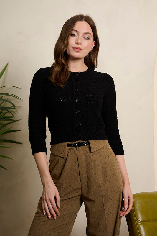 Woman wearing a black sweater and brown pants against a beige wall.