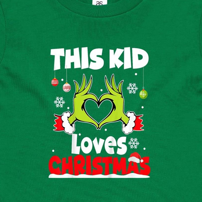 A close-up of a green Christmas t-shirt featuring a graphic of the Grinch's hands forming a heart, with the text 'THIS KID Loves CHRISTMAS' in white and red.