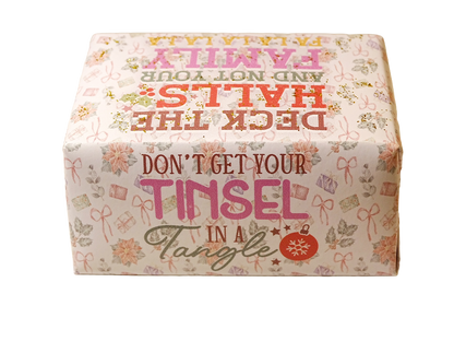 A square decorative box with a festive holiday design, featuring the text 'DON'T GET YOUR TINSEL IN A TANGLE' and 'DECK THE HALLS AND NOT YOUR FAMILY' in pink, red, and green lettering on a cream background with floral and gift motifs.