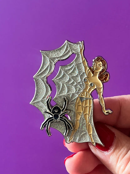 A hand with red nail polish holds an enamel pin featuring a silver spiderweb and black spider design next to a gold woman with red hair, set against a solid purple background.