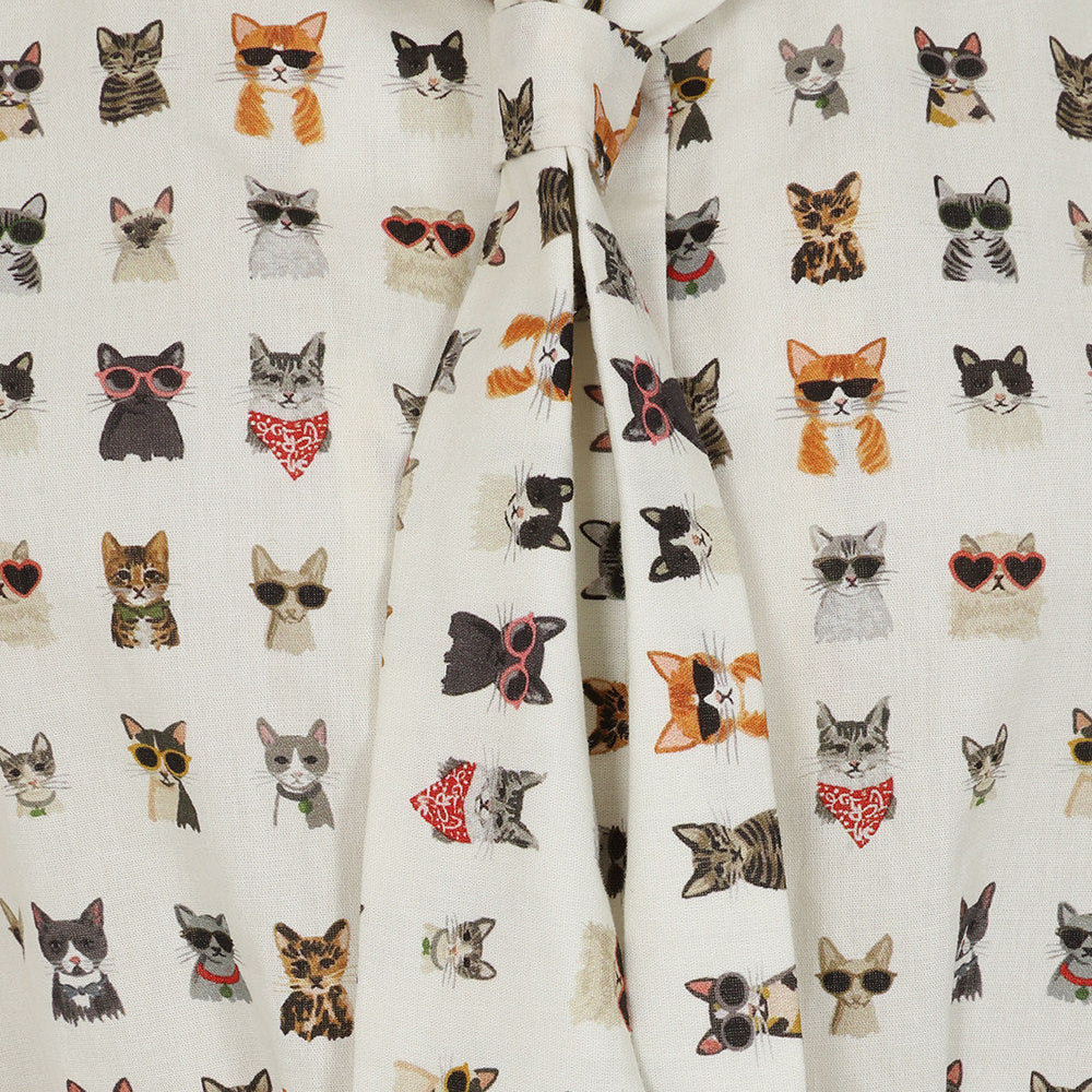 A close-up of a cream-colored fabric garment, likely a tie-front shirt or blouse, featuring an all-over print of various cat faces wearing sunglasses, bandanas, and other accessories.