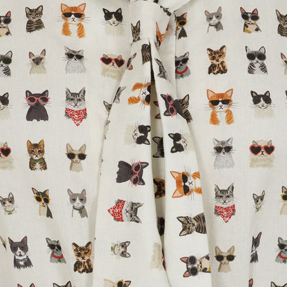 A close-up of a cream-colored fabric garment, likely a tie-front shirt or blouse, featuring an all-over print of various cat faces wearing sunglasses, bandanas, and other accessories.