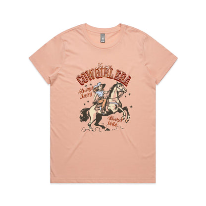 A white graphic t-shirt featuring a retro western illustration of a cowgirl riding a horse. The text reads 'In my COWGIRL ERA' in arched red letters, 'Always Sassy' in red script, and 'Always Wild' in red script, with the cowgirl wearing a cowboy hat and boots while riding a rearing horse.