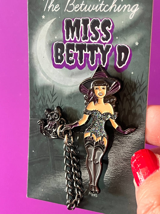 A witch-themed enamel pin featuring a stylized woman in a pointed hat and dress holding a broom, attached to a black chain and displayed on a printed backing card. A hand with red nail polish is holding the card at the right edge against a bright pink background.