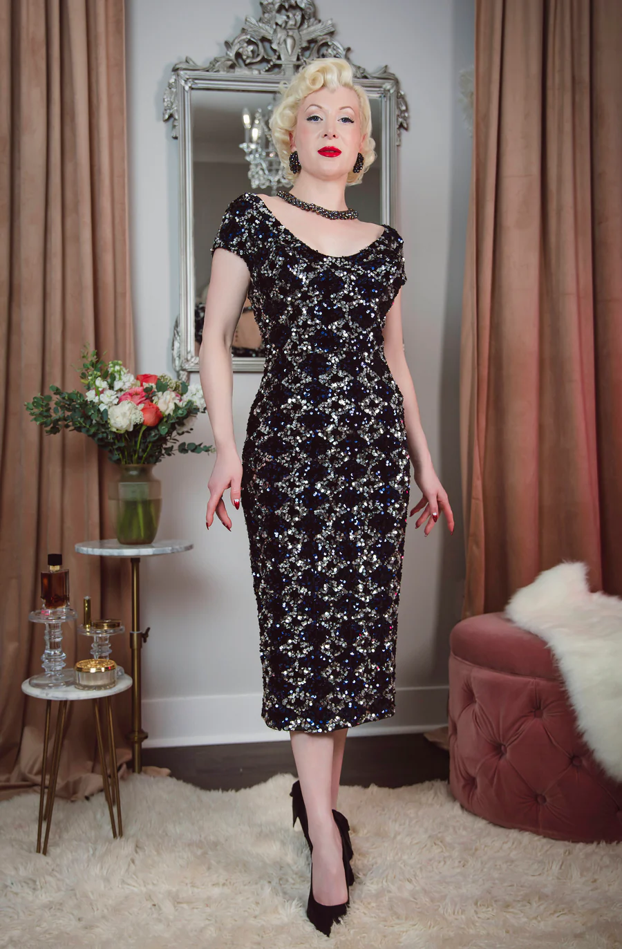 Woman in a black floral dress standing in a room with curtains and a mirror.