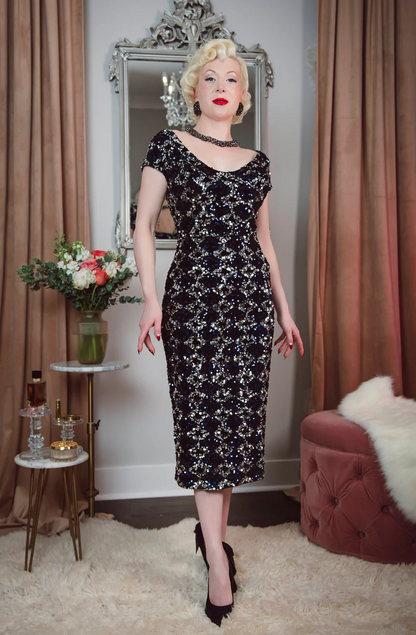 Woman in a black floral dress standing in a room with curtains and a mirror.