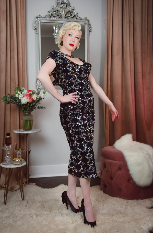 Woman in a black floral dress posing in a room with curtains and a mirror.