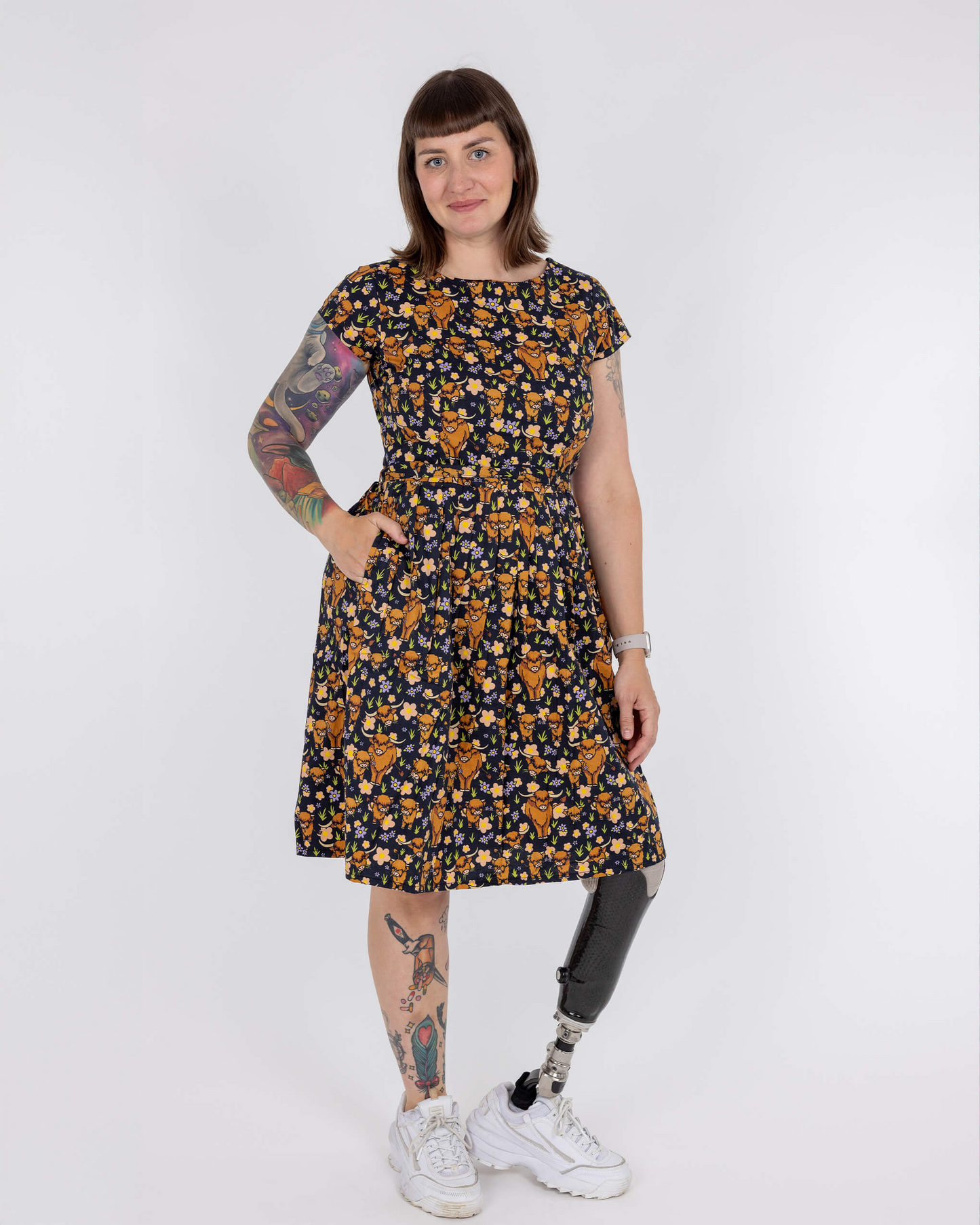 Woman wearing a patterned dress with tattoos on arms and legs, standing against a white background