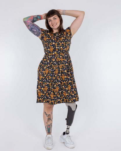 Person wearing a floral dress with tattoos on arms and legs, standing against a white background