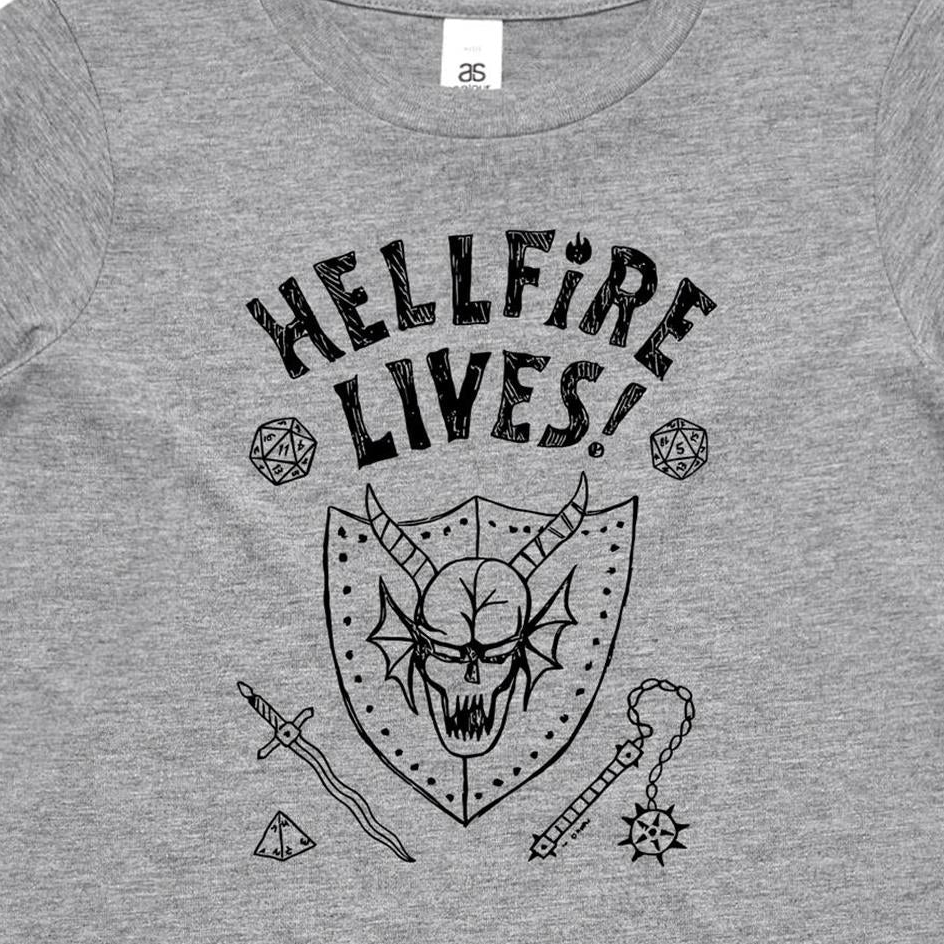 A close-up of a heather gray t-shirt featuring a black graphic print of a horned skull in a shield, surrounded by fantasy-themed icons including dice, a sword, and a mace, with the text 'HELLFIRE LIVES!'.