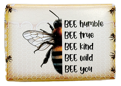 A rectangular decorative plaque or sign featuring a large honeybee illustration over a honeycomb pattern, with the phrase 'BEE humble BEE true BEE kind BEE wild BEE you' printed in black text on the right. The design has a warm beige and yellow background with small bee motifs along the border.